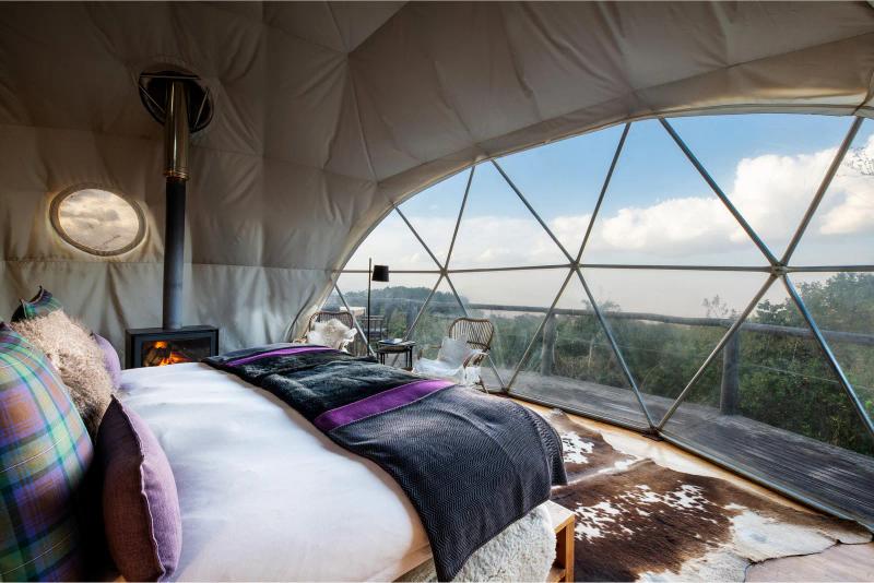 Interior of a domed suite at The Highlands in Tanzania, featuring a bed, fireplace and panoramic windows overlooking the forested landscape.
