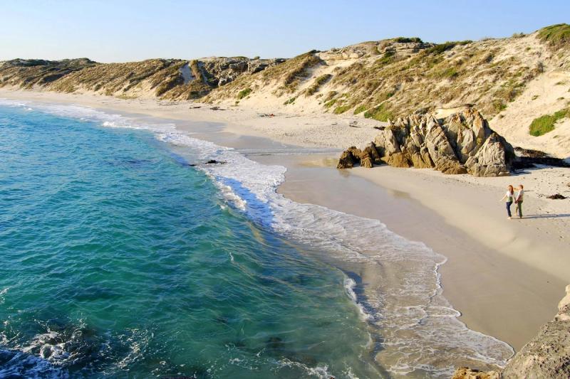 A sweeping view of a secluded, sandy beach at Grootbos Private Nature Reserve, bordered by rolling dunes and turquoise waves. In the lower right corner, a couple walks close to the water’s edge, passing rugged rock formations along the shore.