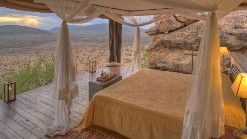An elegant open-air bedroom with a four-poster bed draped in soft white netting, set on a wooden deck with lanterns, overlooking a vast savannah landscape surrounded by rocky outcrops