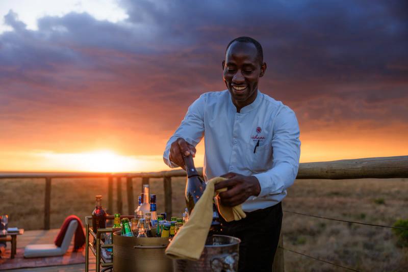 A smiling camp staff member pours a drink from a bottle on a wooden deck at sunset, with drinks and a glowing orange sky behind him.