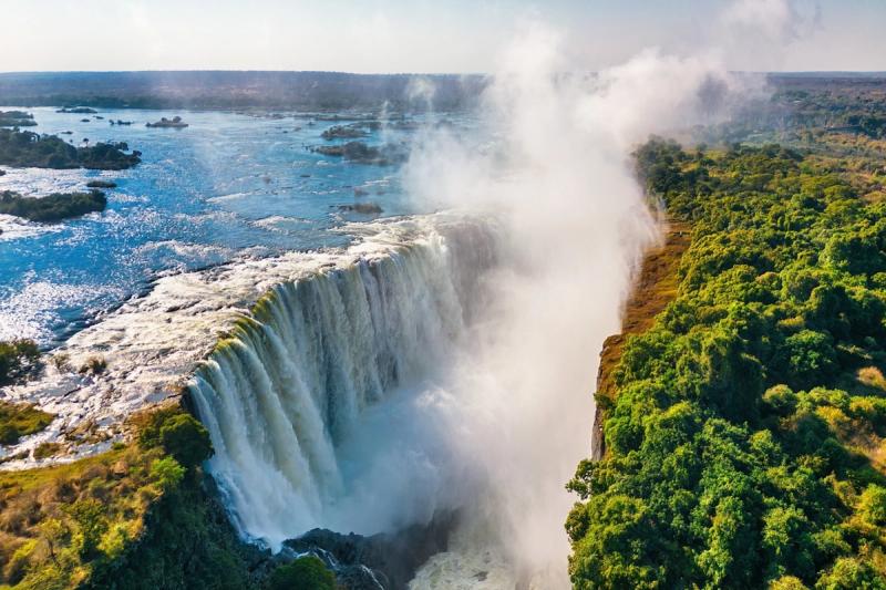 A massive waterfall plunges into a gorge, surrounded by mist and lush green forest, with a wide river flowing above the falls
