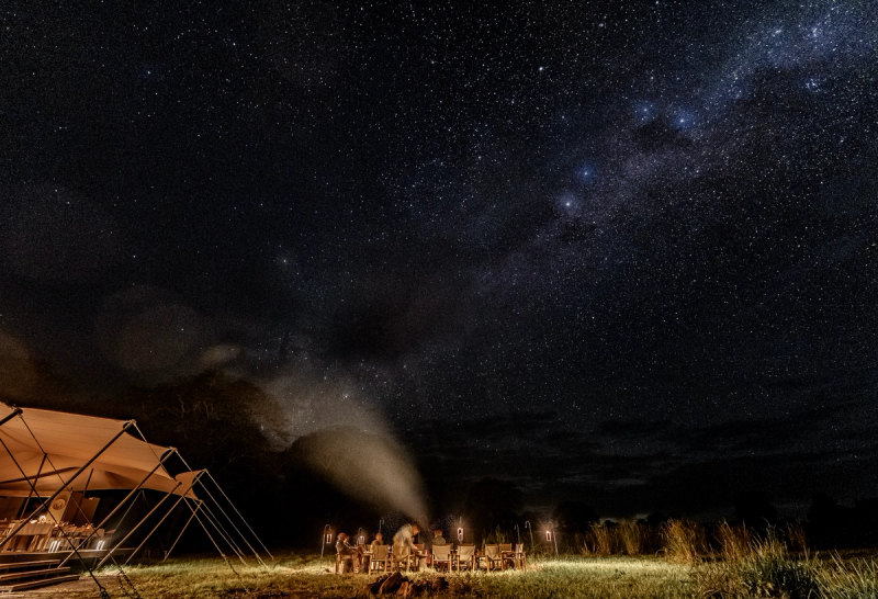 An outdoor safari dinner beneath a star-filled African sky, with guests gathered around a fire beside a luxury tented camp.