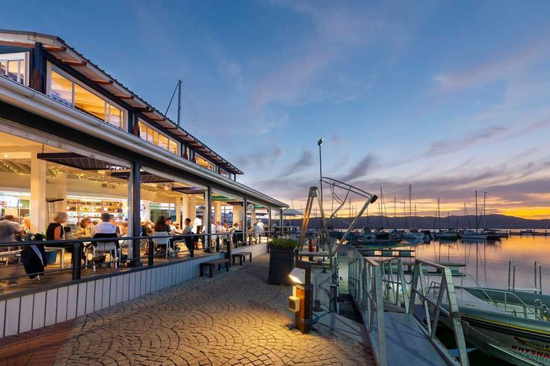 People dine at an open-air waterfront restaurant overlooking a marina at dusk in Knysna