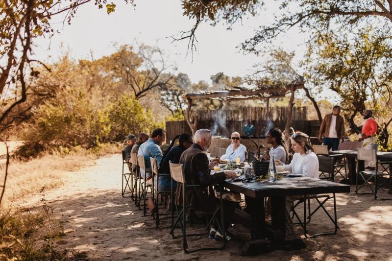 Guests dining at long tables outdoors under trees in a natural bush setting with rustic structures in background