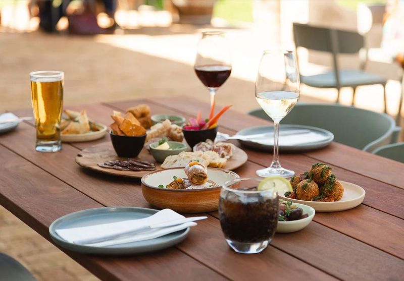 Wooden outdoor table set with wine glasses, beer, soda, and assorted appetizer plates