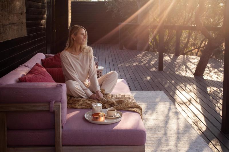 A woman sitting on a pink couch in the sun on a private deck at Silvan Safari with a tea tray beside her, looking out over the bush