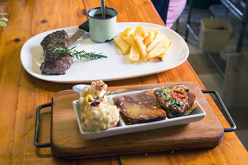 A grilled steak with rosemary, fries, and sauce served beside a wooden board of roasted vegetables and meat
