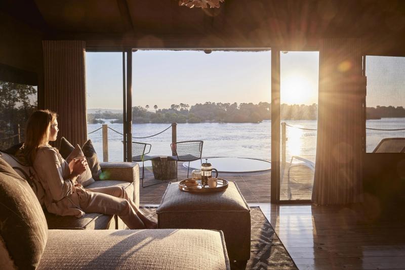 A woman sits with a warm drink, watching the sun rise over a river