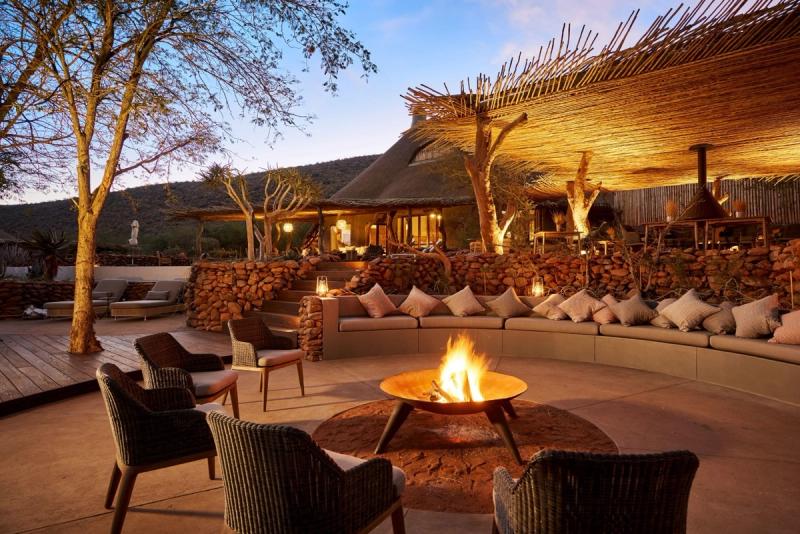 A luxurious outdoor boma seating area, featuring a crackling firepit surrounded by plush cushions and wicker chairs, with warm lantern light illuminating the rustic yet elegant setting under the open sky.