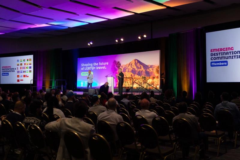 A speaker addresses a seated audience at the IGLTA convention, with a colourful stage backdrop reading “Shaping the future of LGBTQ+ travel.”