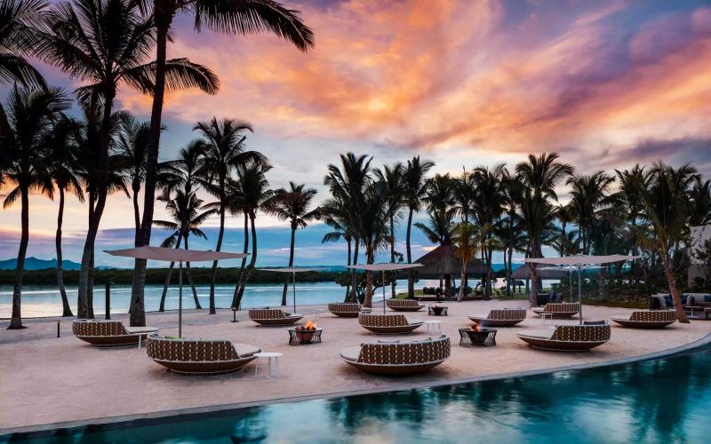 A luxurious beachfront resort glows under a fiery sunset, with stylish sun loungers, flickering fire pits, and swaying palm trees framing the tranquil lagoon.