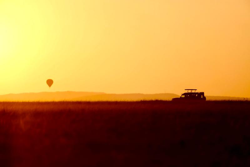 Golden sunrise silhouette of a safari vehicle and a distant hot-air balloon across the vast Maasai Mara horizon.