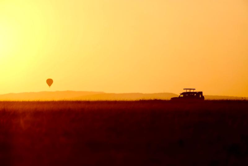 Golden sunrise silhouette of a safari vehicle and a distant hot-air balloon across the vast Maasai Mara horizon.