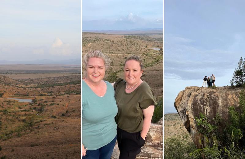A woman stands on a rocky outcrop overlooking a wide, dry landscape of hills, vegetation and distant water, while three people stand on a larger rock nearby.