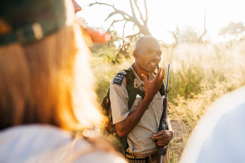 Smiling safari guide holding a rifle speaks with guests in the African bush at golden hour