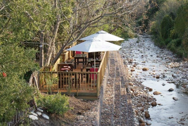 Wooden deck with umbrellas and dining tables beside a rocky river in Swellendam