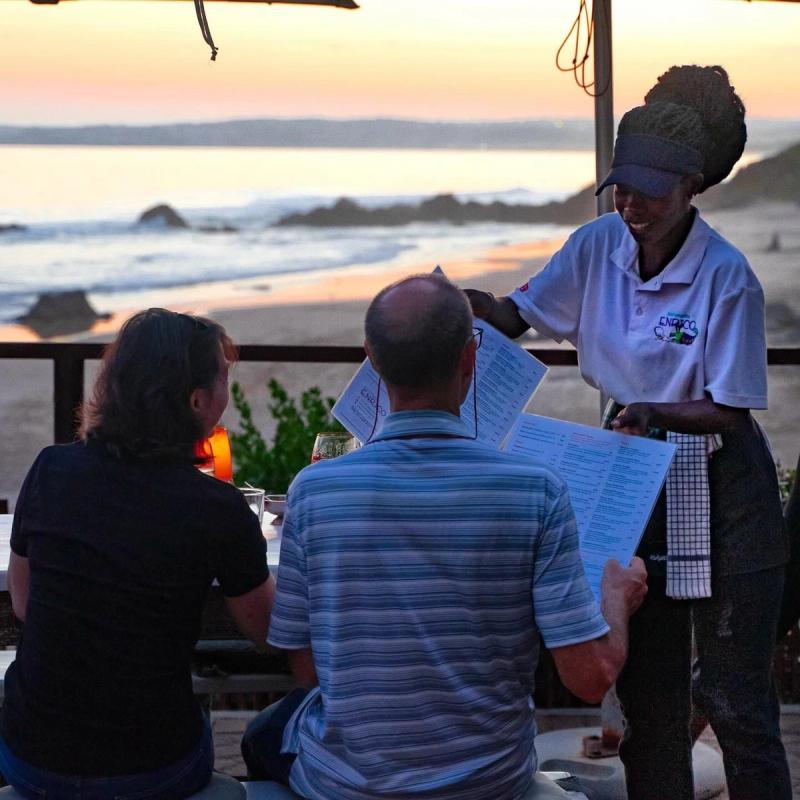 A waitress hands menus to guests seated at a beachfront table during sunset in Plettenberg Bay
