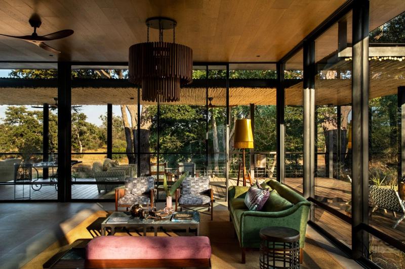 Modern lounge with floor-to-ceiling windows overlooking African bushland, featuring velvet furniture and natural thatched ceiling