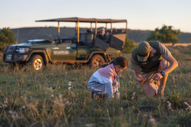  A young girl and a safari guide crouch together in the grassy plains of a malaria-free game reserve, inspecting a small find on the ground with focused curiosity.