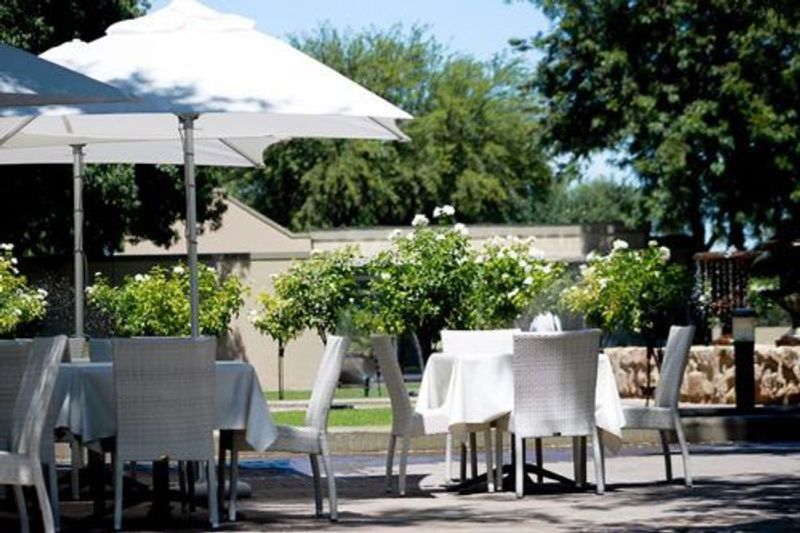 White umbrellas shade table settings on an outdoor patio surrounded by flowering shrubs and trees