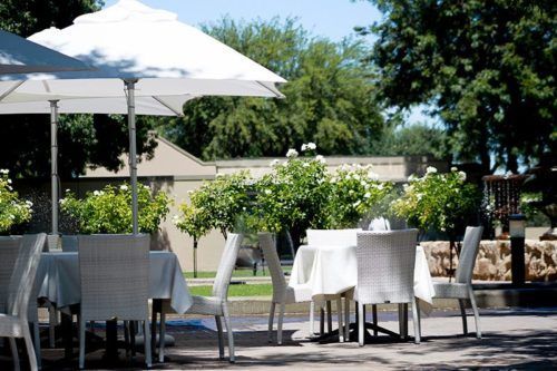 White umbrellas shade table settings on an outdoor patio surrounded by flowering shrubs and trees