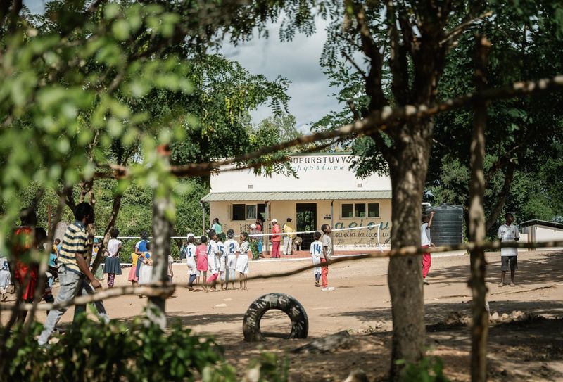 Children gather and play outside a rural school building shaded by trees, showing how education and everyday life are strengthened through the impact in Gorongosa.