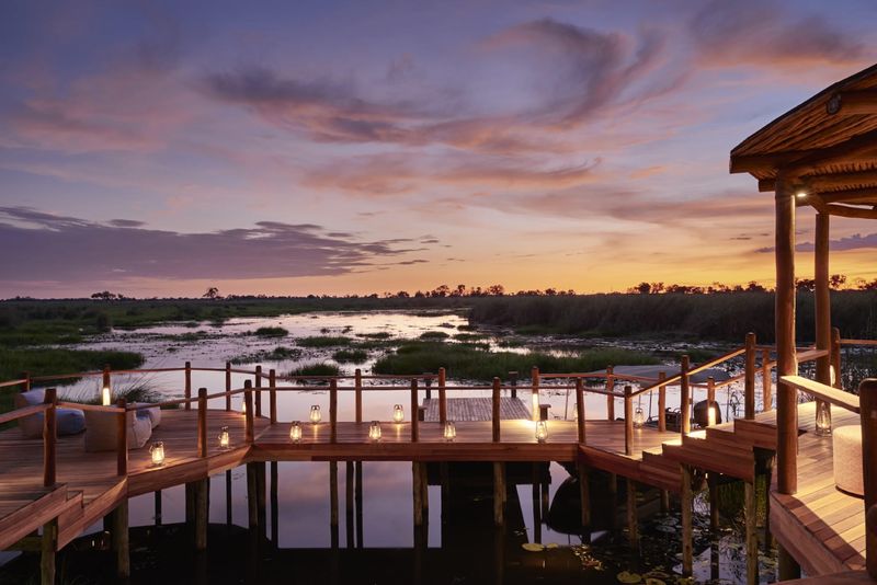A wooden deck with lanterns overlooks a peaceful wetland, bathed in soft purple and orange hues of sunset