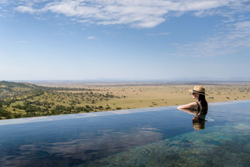 A woman in a straw hat relaxes in an infinity pool overlooking the vast Serengeti plains, with scattered acacia trees and distant hills under a clear blue sky.