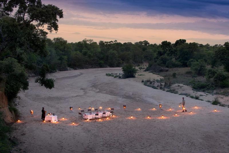 A romantic lantern-lit dinner set up on a sandy dry riverbed at dusk surrounded by trees