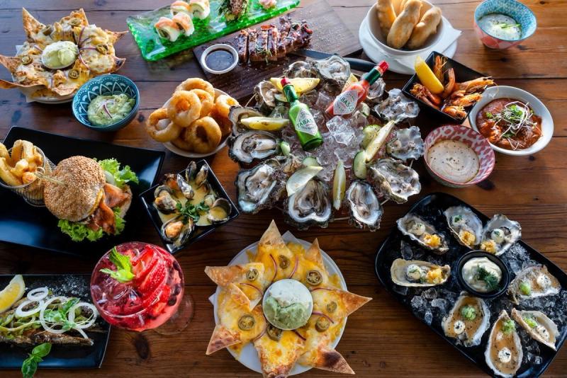 Overhead view of a wooden table filled with oysters, burgers, nachos, and seafood dishes