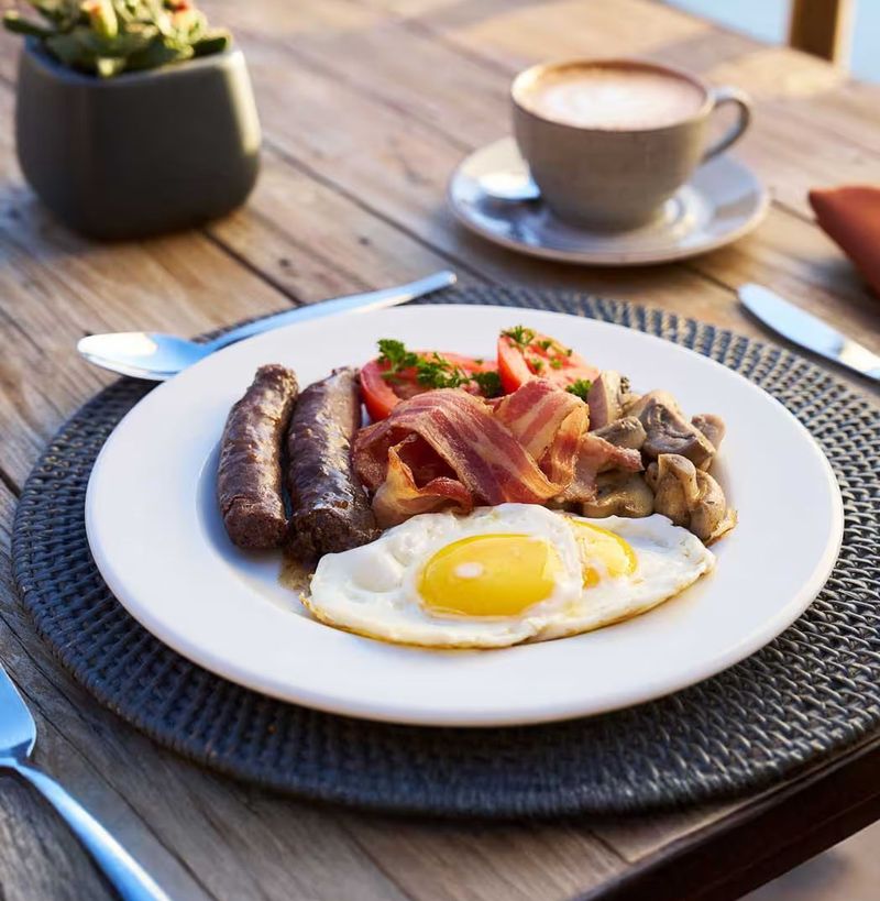Breakfast plate with eggs, sausages, bacon, mushrooms and tomato served outdoors with coffee