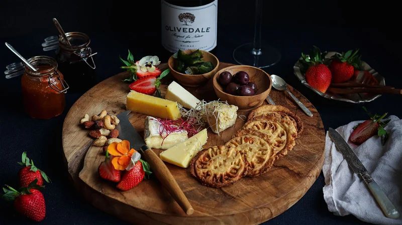 Wooden board with cheeses, crackers, nuts, strawberries and olives beside a wine bottle