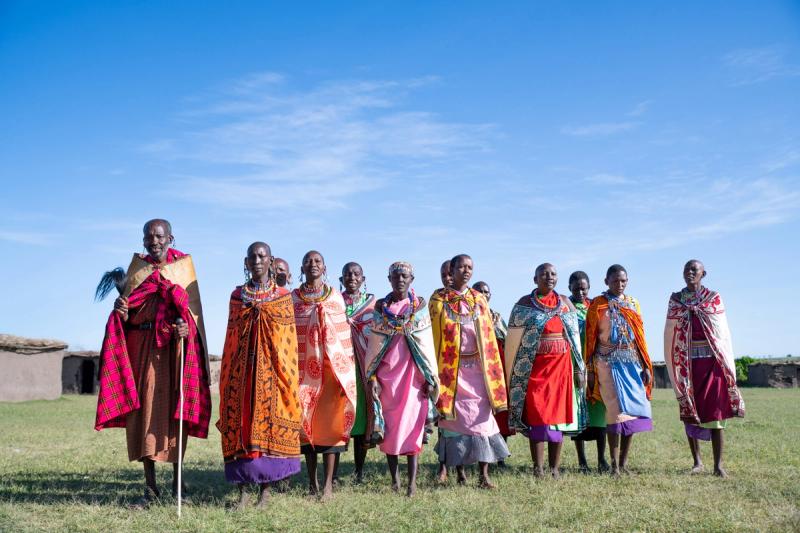 A group of Maasai people standing in a line on grassy ground, wearing vibrant, patterned clothing and ornate beadwork, against a clear blue sky with traditional huts in the background.