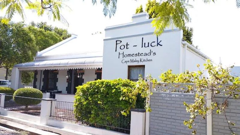 White gabled building with a sign reading Pot-luck Homestead's Cape Malay Kitchen, surrounded by greenery