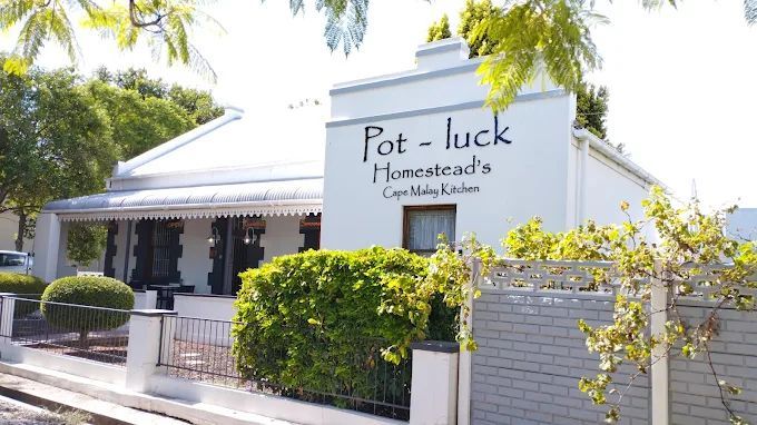 White gabled building with a sign reading Pot-luck Homestead's Cape Malay Kitchen, surrounded by greenery