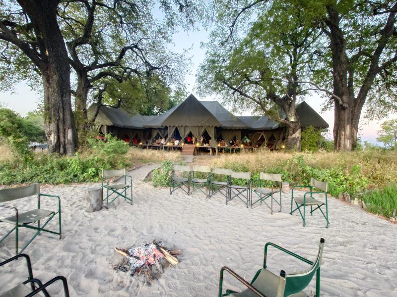 A safari camp dining and lounge area opens onto a sandy clearing with canvas chairs arranged around a central fire pit.