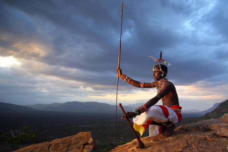 A Samburu warrior in traditional dress kneels on a rocky outcrop at sunset, overlooking vast wilderness – a powerful visual to end your search for the best time to visit Kenya.