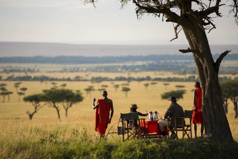A small group enjoys an outdoor meal under a tree, with Maasai guides nearby and vast savannah stretching into the distance