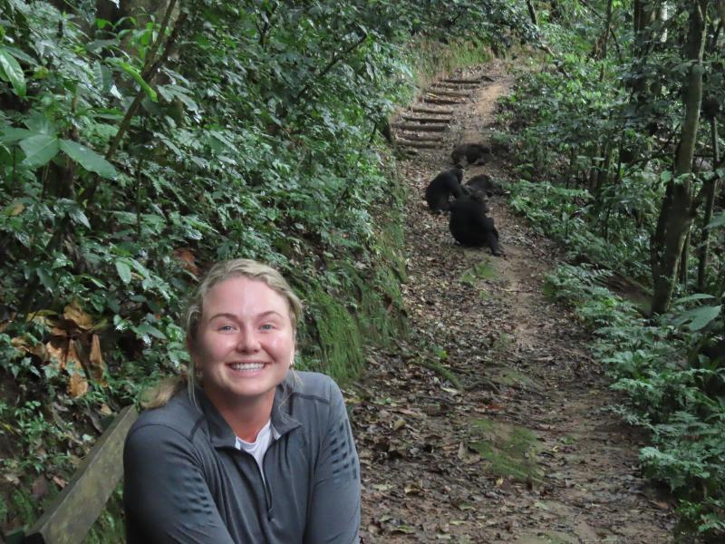Janine smiles for a photograph on a forest trail during a gorilla trek, with gorillas visible further along the path.