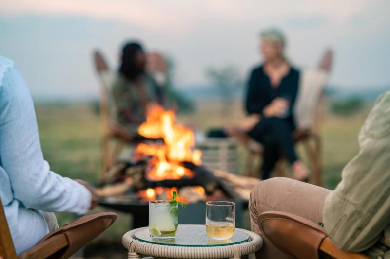 A cosy campfire with drinks on a table, surrounded by blurred figures seated in the open savannah