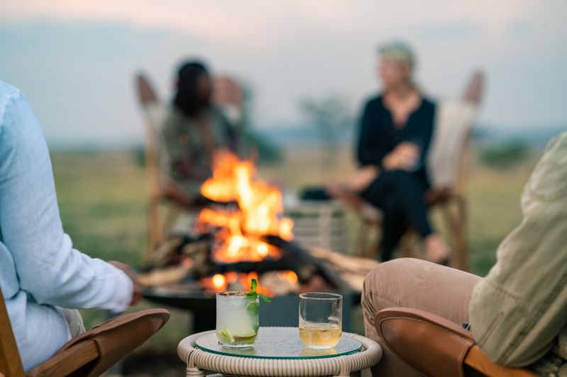 A cosy campfire with drinks on a table, surrounded by blurred figures seated in the open savannah