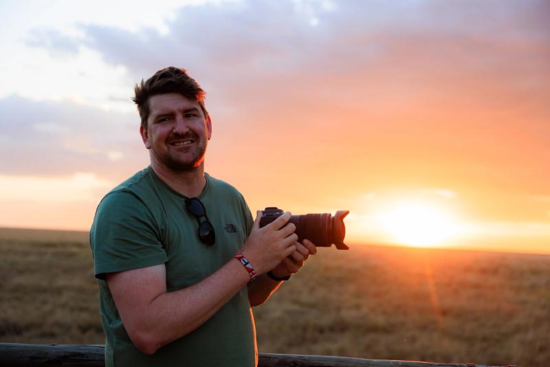 A smiling man holds a camera with a telephoto lens against a warm sunset over the Maasai Mara savannah.