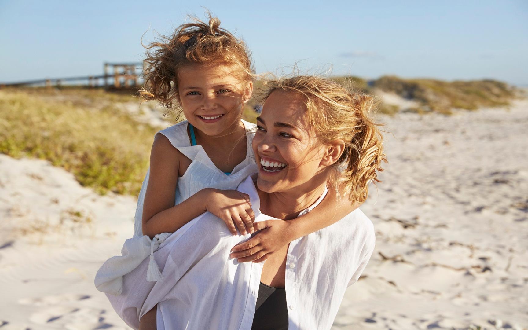 Bildet viser en voksen og et barn som er på en strand. Den voksne bærer barnet på ryggen, og begge smiler bredt.