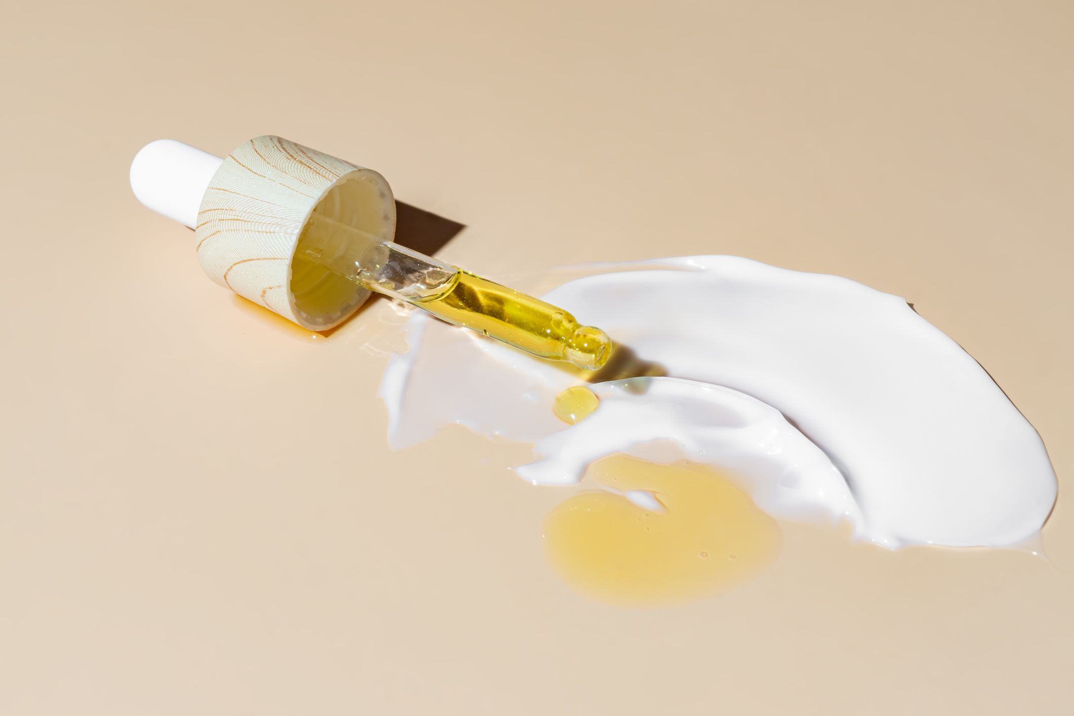 Nærbilde av en pipette med gyllent ansiktsserum plassert ved siden av en hvit hudkrem på en lys, nøytral bakgrunn.