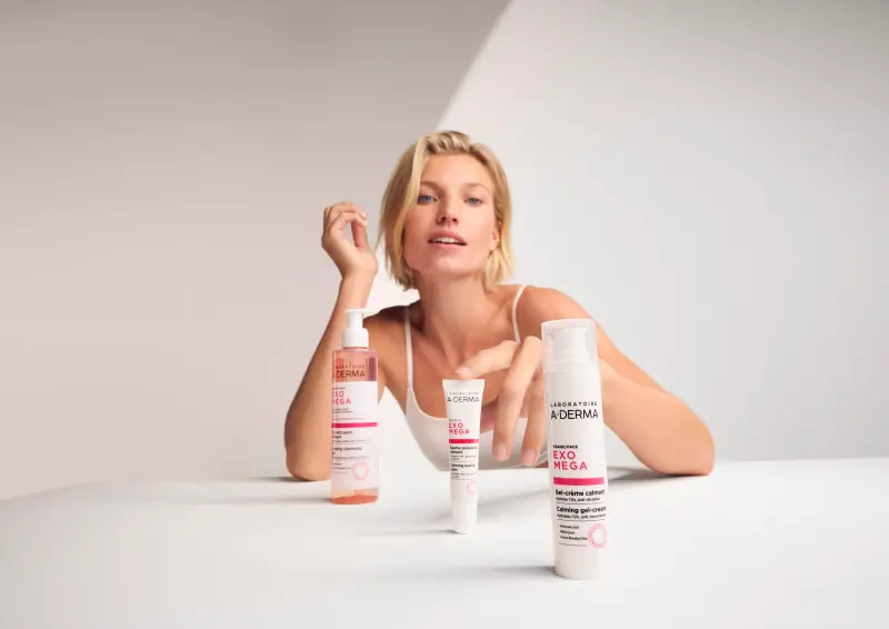 Kvinne sitter ved et bord med produkter fra A-derma Exomega ansiktserien fremfor seg