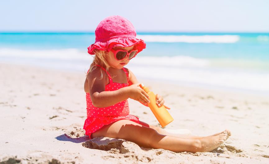 En liten jente med rosa badedrakt og solhatt sitter på stranden med en solkrem i hendene