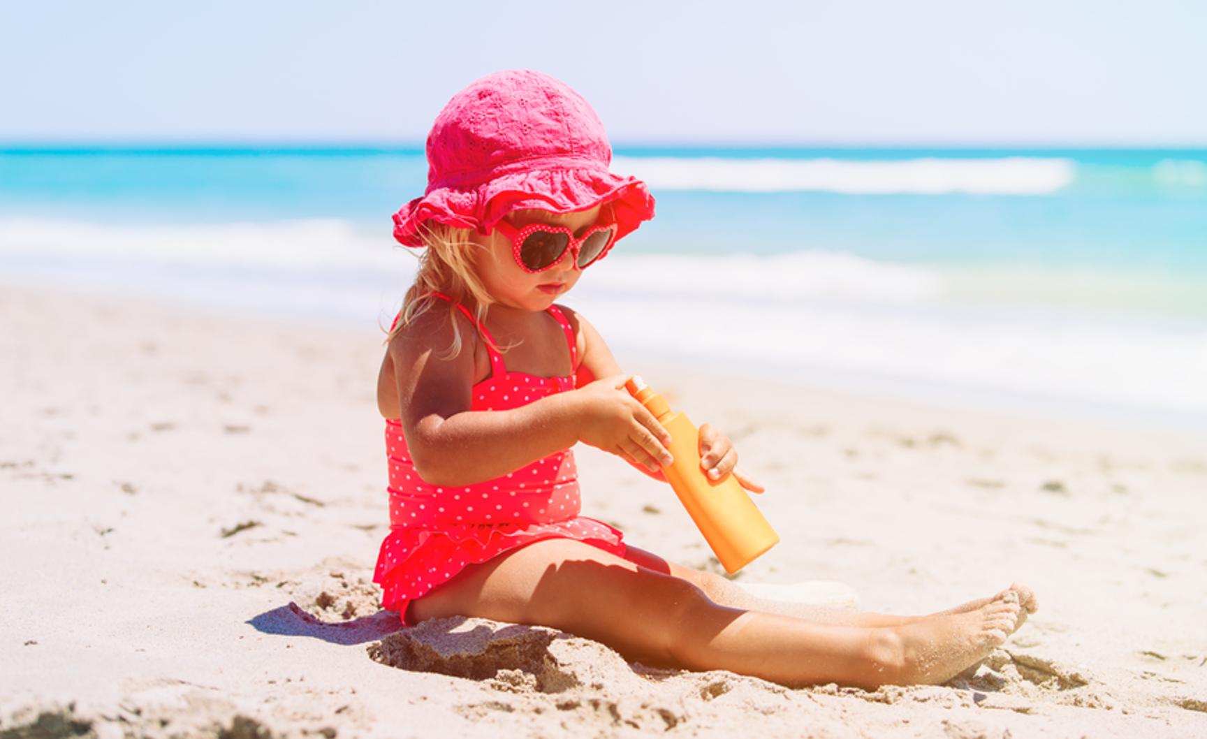 En liten jente med rosa badedrakt og solhatt sitter på stranden med en solkrem i hendene