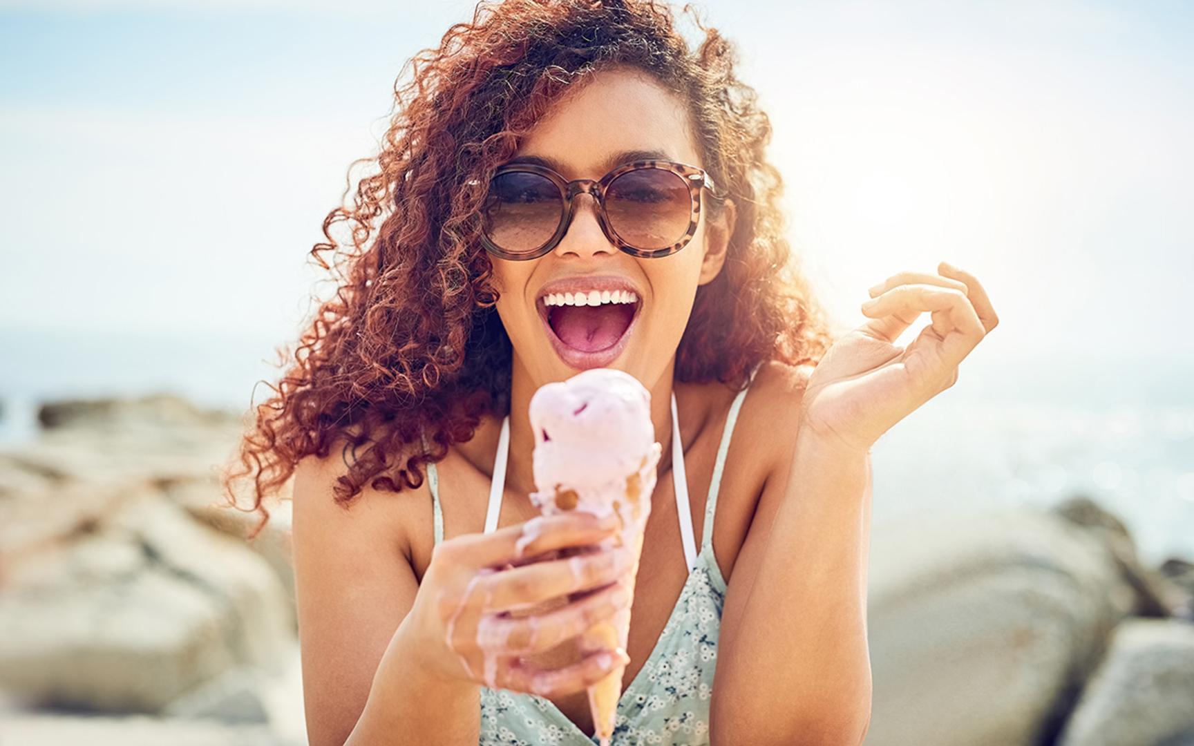 Dame med krøller og solbriller spiser is sittende ved sjøen