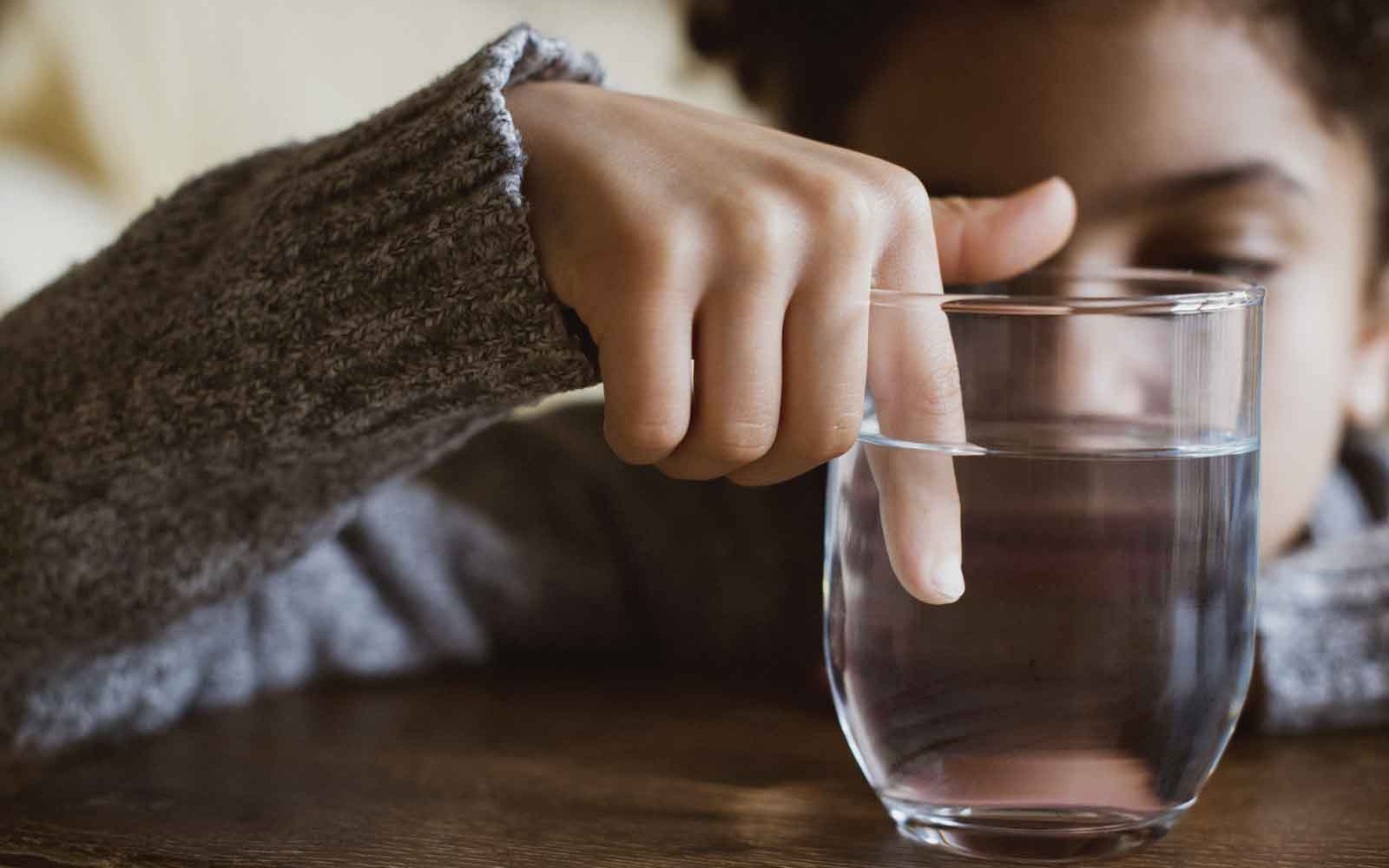 Et barn sitter ved et bord og holder pekefingeren i et glass med vann