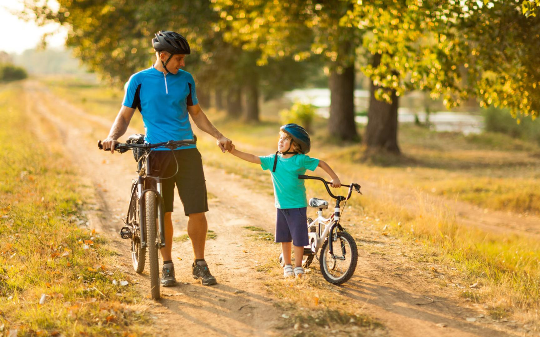 En mann og en gutt leier hver sin sykkel langs en grusvei blant trær.